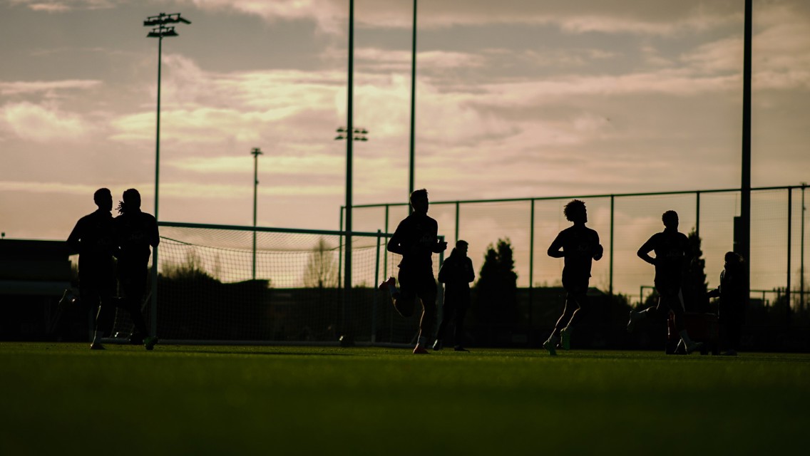 Silhouetted players running on a soccer field during sunset with goalposts and tall lights in the background.