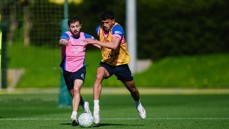 Two football players in training, one wearing a pink bib and the other in a yellow bib, both competing for a ball during a practice session on a grass field.