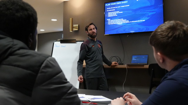Three people are in a meeting room, a person is presenting a 'Plan Your Own Event' session with instructions visible on a screen. A whiteboard with the title 'Desert Island' stands in the background.