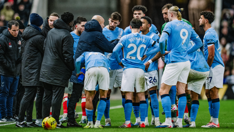 A group of Manchester City players and coaches huddle together on the football pitch during a match. They are wearing light blue jerseys with visible names and numbers.