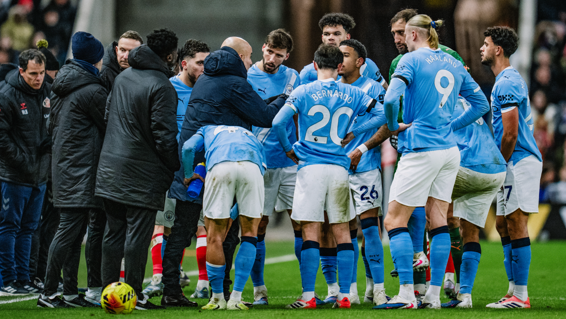 A group of Manchester City players and coaches huddle together on the football pitch during a match. They are wearing light blue jerseys with visible names and numbers.