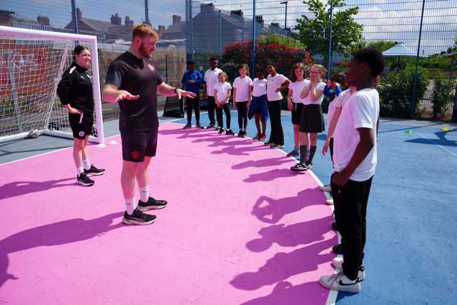 A man is seen teaching a group of young kids on an outdoor football pitch. The children are standing in line as they listen to the man who appears to be giving instructions.