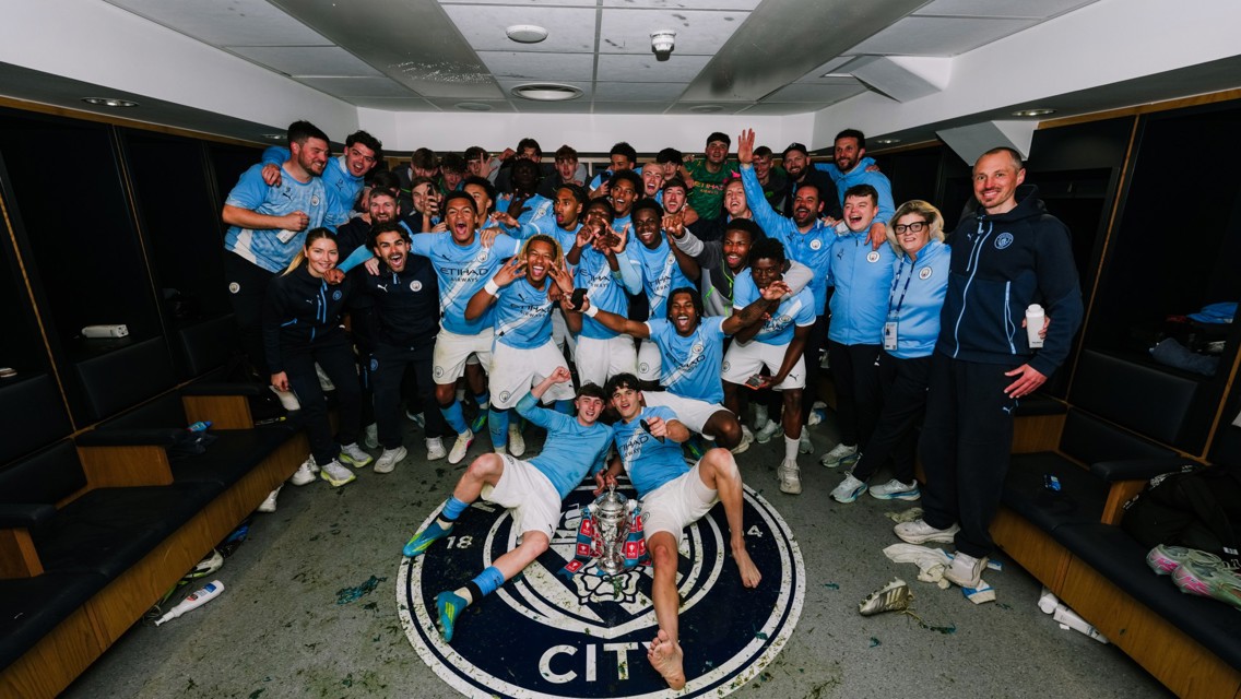Group of football players and staff celebrating in a locker room with a trophy on the floor, surrounded by a Manchester City emblem.