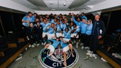 Group of football players and staff celebrating in a locker room with a trophy on the floor, surrounded by a Manchester City emblem.