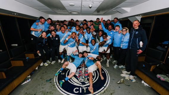 Group of football players and staff celebrating in a locker room with a trophy on the floor, surrounded by a Manchester City emblem.
