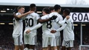 Fulham players in white jerseys celebrating a goal with a group hug on the field. The crowd and a scoreboard with 'GOA' visible in the background.