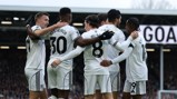 Fulham players in white jerseys celebrating a goal with a group hug on the field. The crowd and a scoreboard with 'GOA' visible in the background.