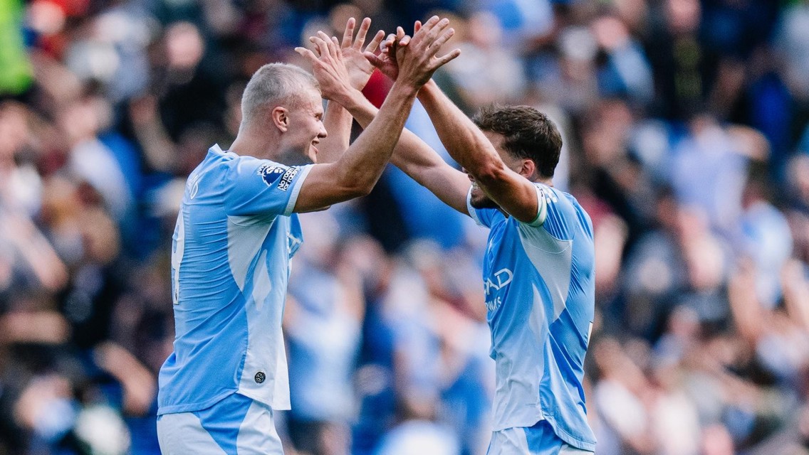Two Manchester City players in blue kits celebrate together on the field, raising their hands for a high-five, with a blurred crowd in the background.