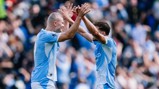 Two Manchester City players in blue kits celebrate together on the field, raising their hands for a high-five, with a blurred crowd in the background.
