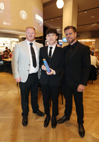 Three men dressed formally standing together indoors. One in the center is holding an award. The background shows people seated and a projected logo on the wall.