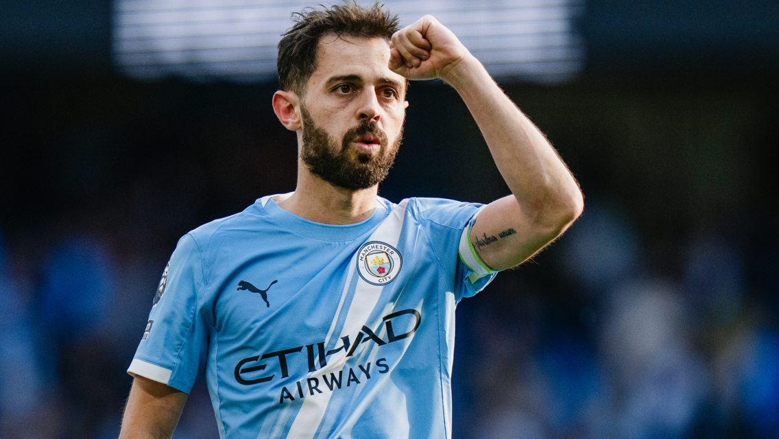 The image shows a football player wearing a Manchester City kit and celebrating with a clenched fist.
