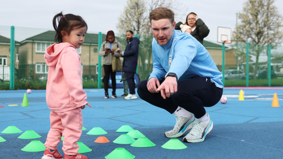 A coach is squatting and pointing while talking to a young child standing near cones on an outdoor sports court, with a group of adults in the background.