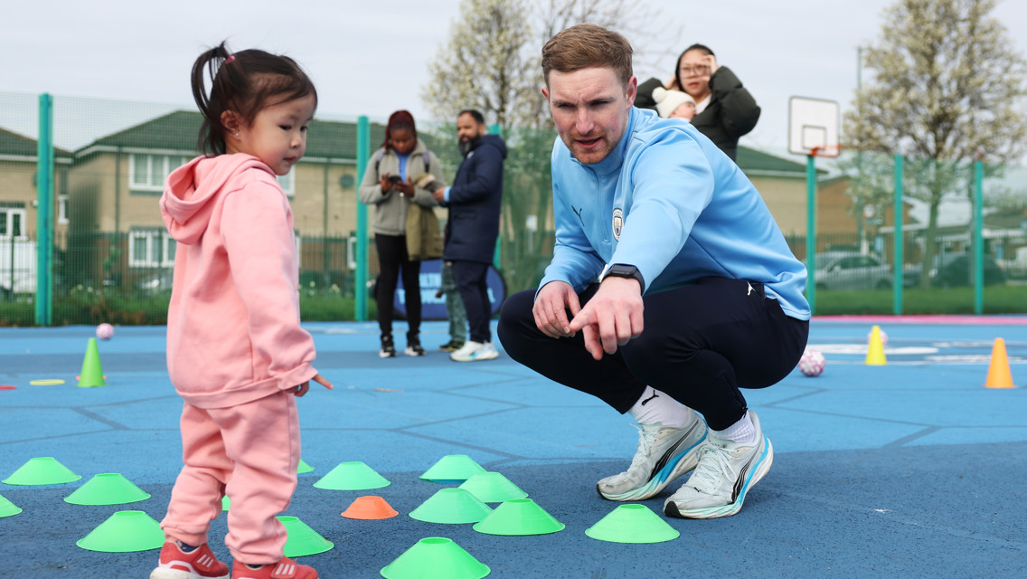 A coach is squatting and pointing while talking to a young child standing near cones on an outdoor sports court, with a group of adults in the background.