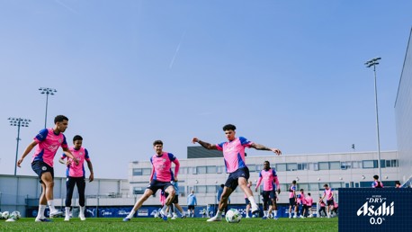 A group of players in pink training bibs practicing on a grassy field with footballs around, buildings and floodlights in the background. The scene features a blue sky and an Asahi sponsor logo.