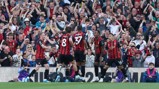 Football team players in red and black kits celebrate in front of cheering crowd.