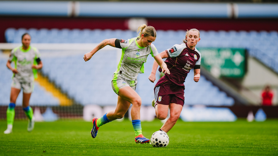Two female soccer players in opposing uniforms compete for the ball on a stadium field during a match.