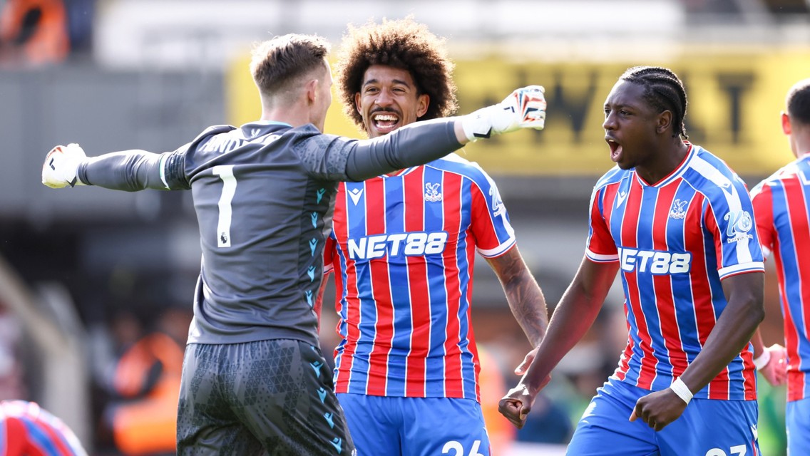 Three football players celebrate on the field in colorful uniforms, one wears a grey goalkeeper outfit with the number 1. They appear joyful and energetic.