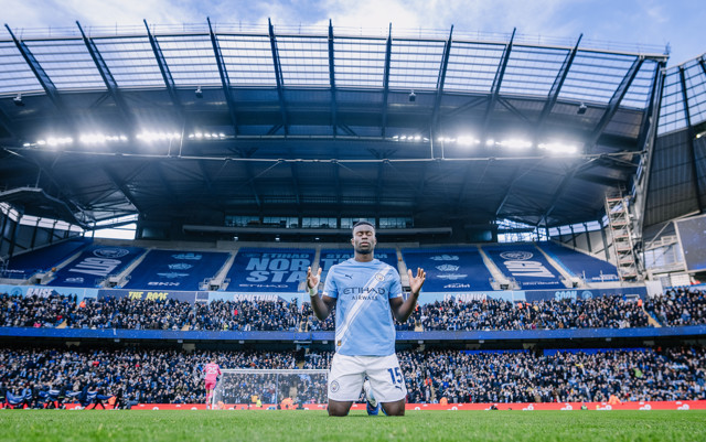 A football player in a Manchester City kit kneels with arms raised in celebration at Etihad Stadium. The stadium is filled with fans, and prominent 'Etihad' and 'North Stand' signage are visible.