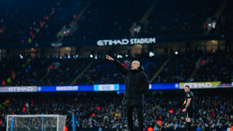 A football coach in a black jacket gestures from the sidelines during a match at Etihad Stadium, with a referee nearby and a full crowd in attendance.