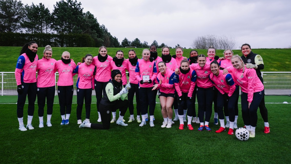 A group photo of the Manchester City women's team wearing pink training gear on a soccer field.