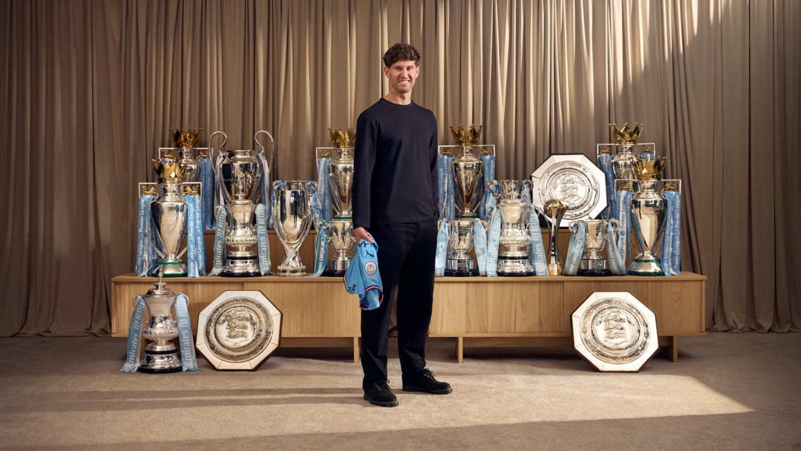 A person stands in front of a collection of football trophies, including Premier League, FA Cup, and Community Shield trophies, associated with Manchester City.
