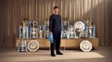 A person stands in front of a collection of football trophies, including Premier League, FA Cup, and Community Shield trophies, associated with Manchester City.