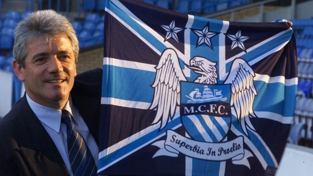 A man holding a Manchester City flag with the club's crest showing 'Superbia in Proelio'.