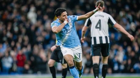 A Manchester City player celebrates a goal with a gesture pointing upward, wearing the team's light blue kit. Newcastle United's Botman is visible in the background wearing a black and white kit.