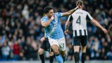 A Manchester City player celebrates a goal with a gesture pointing upward, wearing the team's light blue kit. Newcastle United's Botman is visible in the background wearing a black and white kit.