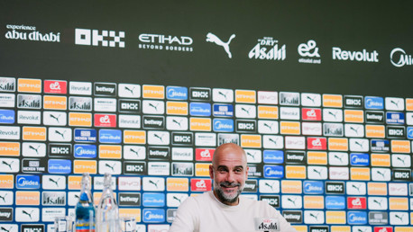 A press conference setting with a person at a table in front of a wall filled with sponsor logos including Etihad, Puma, and OKX. Bottles of water are on the table.