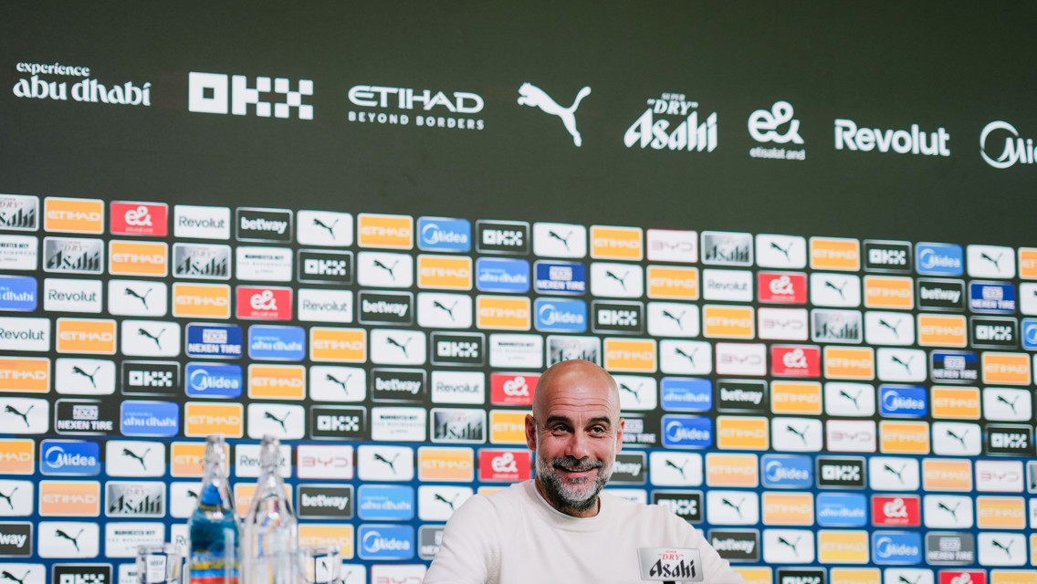 A press conference setting with a person at a table in front of a wall filled with sponsor logos including Etihad, Puma, and OKX. Bottles of water are on the table.