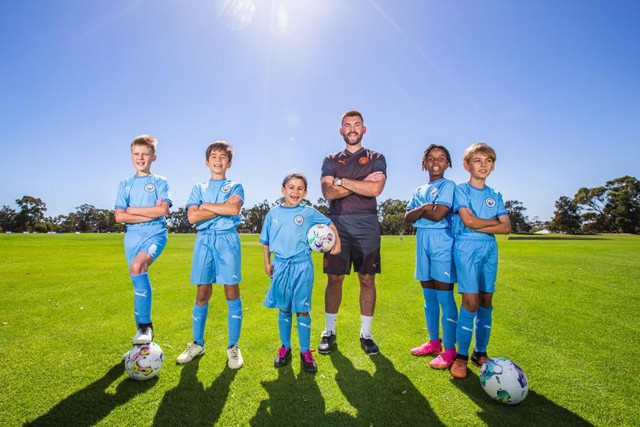 Manchester City football coach with five young players in blue training kits posing on a grassy field on a sunny day.