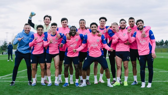 Group of soccer players on a field wearing pink Man City training gear, posing with arms raised in a celebratory fashion.