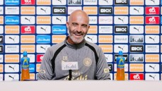 A coach at a press conference with Manchester City logos behind him, two Lucozade bottles in front.