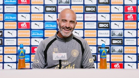 A coach at a press conference with Manchester City logos behind him, two Lucozade bottles in front.