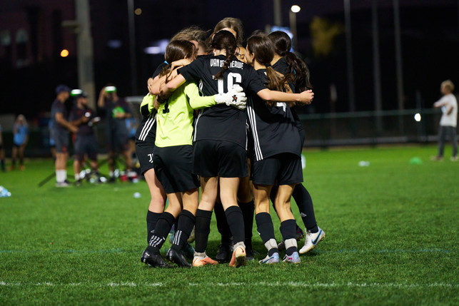 A group of women's soccer players in black uniforms, gathered in a huddle with arms around each other on a grass field, during nighttime.