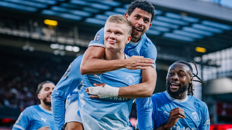 Football players in Manchester City jerseys celebrate a goal together on the field. One player is carrying another on his back, while two others smile joyfully.