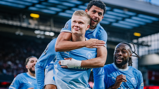 Football players in Manchester City jerseys celebrate a goal together on the field. One player is carrying another on his back, while two others smile joyfully.