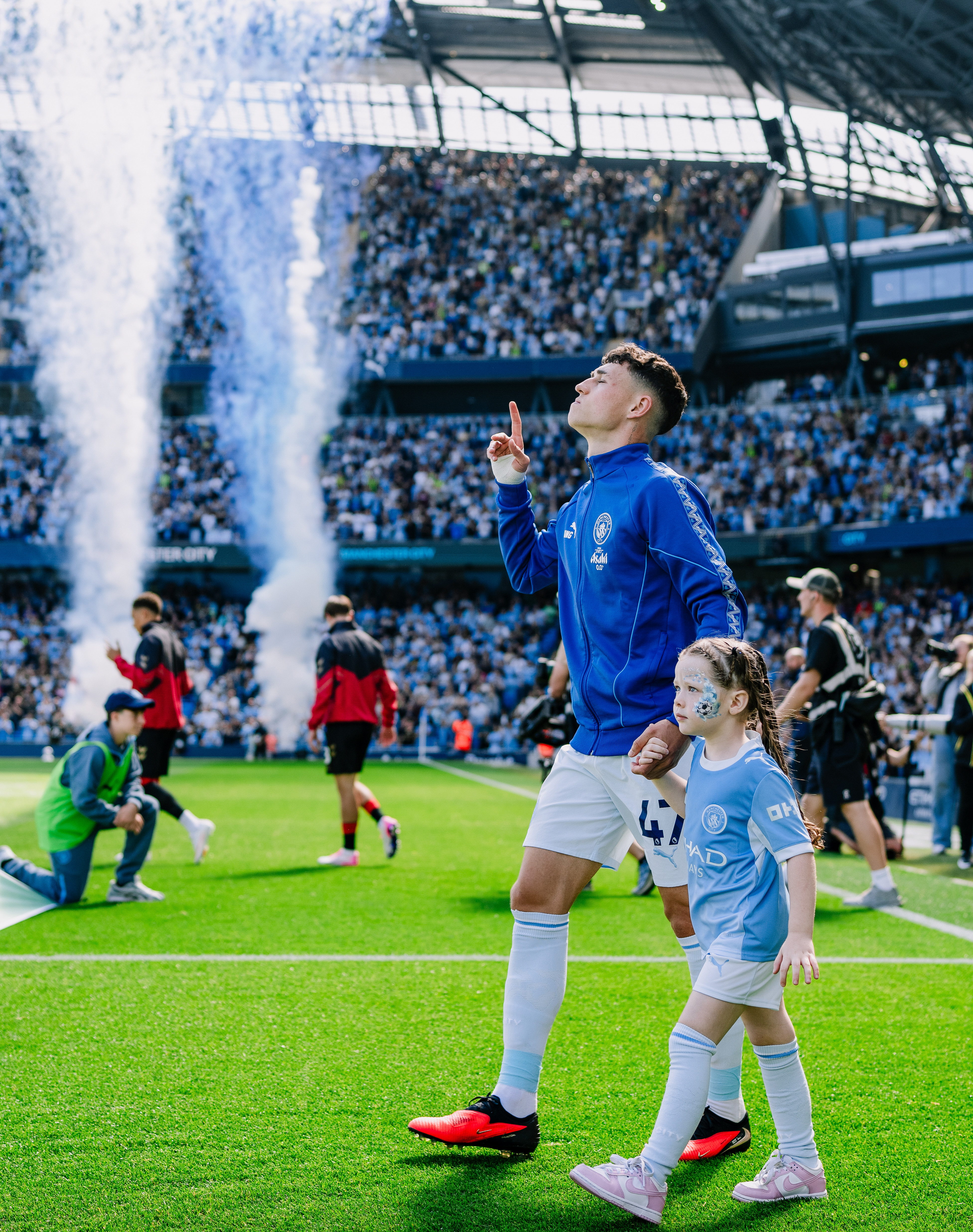A football player walks onto the field at a stadium with a young girl. The child has face paint and wears a Manchester City kit. The stadium is filled with fans, and fireworks are going off.