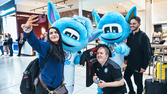 Two mascots, Moonbeam and Moonchester, posing with fans for a selfie at a public location. The mascots are wearing Manchester City attire indicating a celebration of their championship victory.