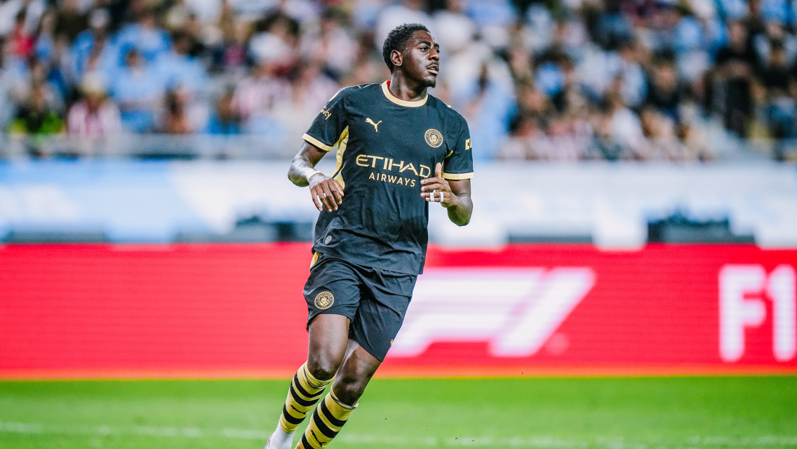 A football player in a Manchester City uniform running on the pitch during a match. The uniform is dark with yellow accents.
