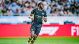 A football player in a Manchester City uniform running on the pitch during a match. The uniform is dark with yellow accents.
