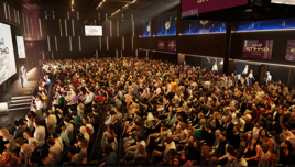 A large audience seated in a hall with Etihad Airways branding on screens. The stage features a presenter in front of the crowd.