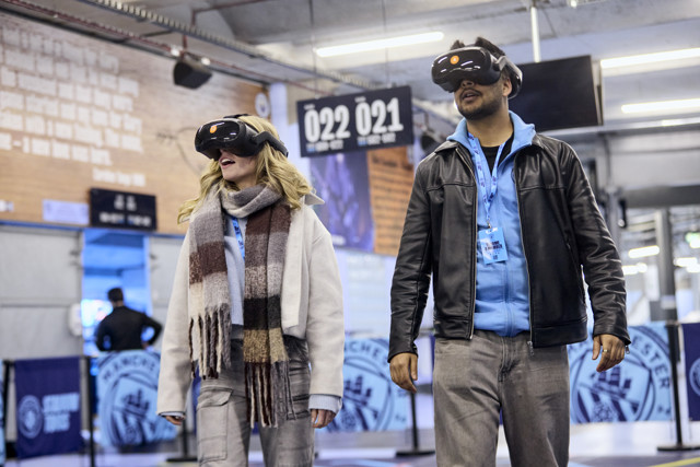 Two people wearing VR headsets and casual clothing inside a stadium-like environment.