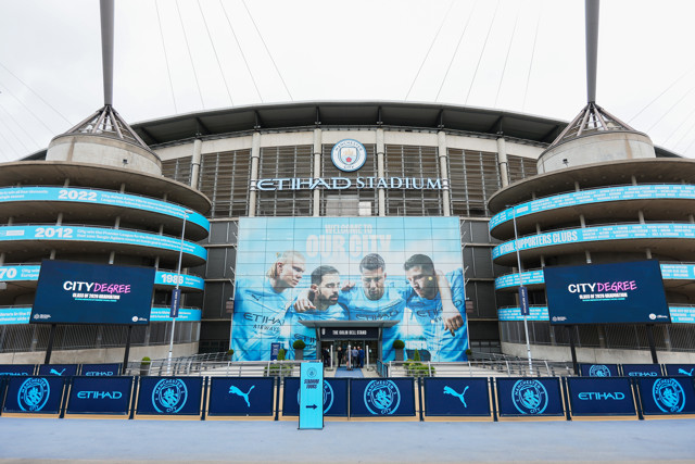 Front view of Etihad Stadium with Manchester City branding and player posters displayed above the entrance.