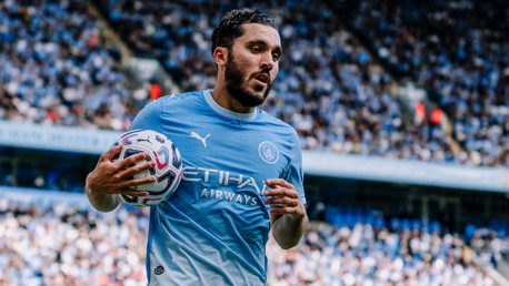 A football player in a Manchester City jersey holding a ball during a match at a crowded stadium.