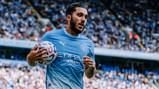 A football player in a Manchester City jersey holding a ball during a match at a crowded stadium.