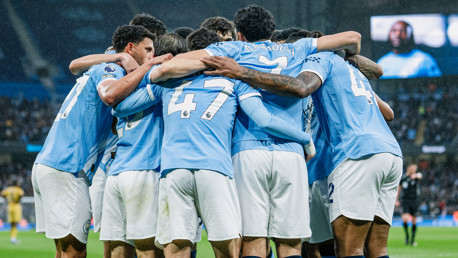 Football players wearing light blue jerseys with white numbers gather in a group huddle on the field celebrating a goal.