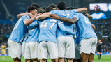 Football players wearing light blue jerseys with white numbers gather in a group huddle on the field celebrating a goal.