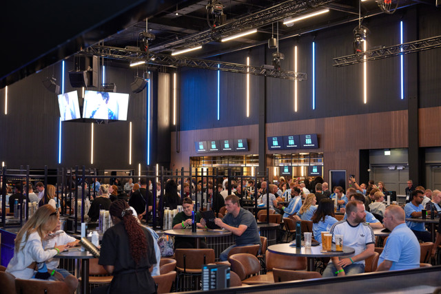A bustling hospitality area at the Etihad Stadium, with fans seated at tables, many wearing light blue jerseys, and a screen overhead displaying a program.